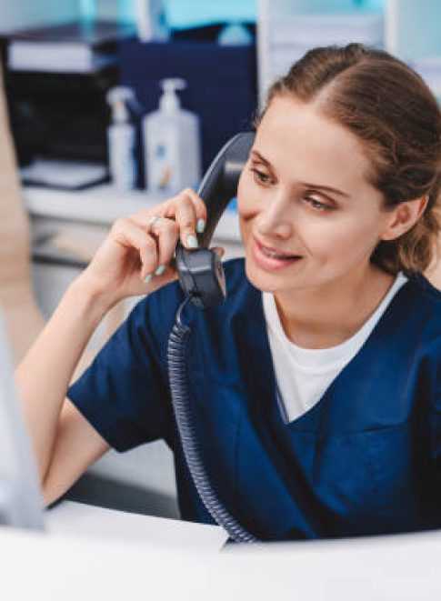 Young female receptionist talking on phone in clinic while sitting and looking on pc monitor